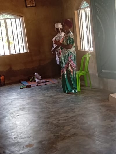 Inside a dimly lit room in Isinyi-Nando, Nigeria, three individuals are present. Two adult women stand in the middle ground, facing left. The woman in the foreground wears a patterned green, pink, and white dress with a red headscarf, her hands clasped in front of her chest. Behind her, another woman wears a white top and a white headscarf, also with hands clasped. On the floor to the left, a person in white clothing lies prone on a blue and white patterned mat, with a green fabric object nearby. Two pairs of sandals (one green, one brown) rest beside the mat. A bright green plastic chair is positioned to the right of the standing women. The room features light brown, textured walls, a grey concrete floor, and two windows with white bars, allowing natural light to enter. The left window is rectangular, while the right window has an arched top. Green foliage is visible outside the windows. A small, dark-framed picture hangs on the upper left wall. A dark, cylindrical container is partially visible on the floor in the far left corner. A dark, ornate doorway is partially visible on the far right.