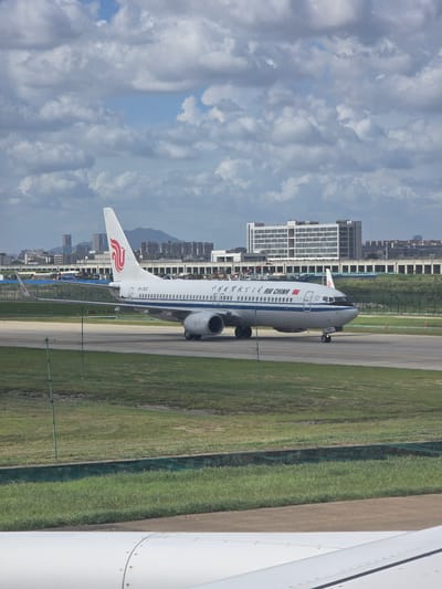 An Air China airplane, with registration B-1763, is on the tarmac at Shenzhen Bao'an International Airport, located in Bao'an District, China. The aircraft is white with blue trim and the Air China logo on its tail. The plane is positioned on a taxiway, with its front landing gear visible. In the background, there are buildings, a fence, and a grassy area. The sky is partly cloudy with blue patches. The image appears to be taken from another aircraft, as a portion of a white airplane wing is visible in the foreground.