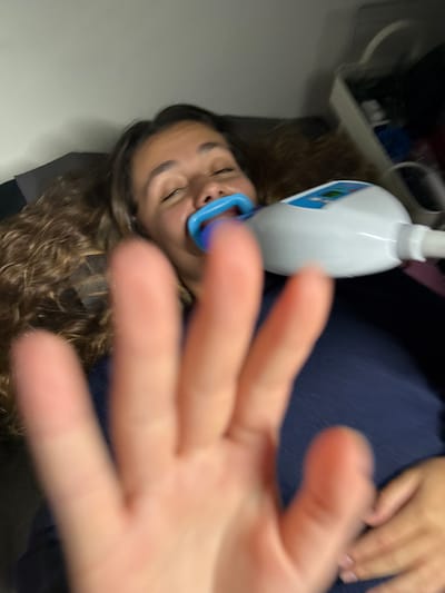 A woman is lying down with a teeth whitening device in her mouth. She has long, curly brown hair and appears to be smiling slightly with her eyes closed. A hand is out of focus in the foreground, partially obscuring the view. The background shows a white wall, and a piece of equipment is visible on the left. The dominant colors are white, blue, and brown.