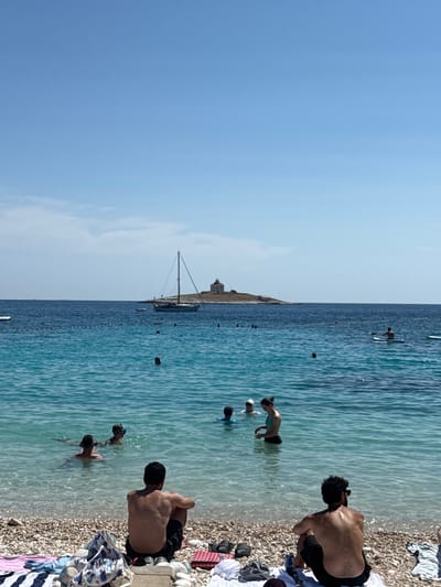 The image depicts a sunny day at a pebble beach in Hvar town, Croatia, with people enjoying the clear turquoise waters of the Adriatic Sea. In the foreground, two men are seated on the beach, facing the sea, with towels and beach bags at their sides. The water is shallow and clear, revealing the pebbles on the seabed. Several individuals are visible in the water, swimming and wading. Further out in the sea, a sailboat is anchored near a small, rocky island topped with a lighthouse or a small building. Other boats, including a paddleboarder, are also present in the distance. The sky is a clear blue with a few wispy clouds. The scene conveys a sense of leisure and relaxation typical of a Mediterranean summer day.