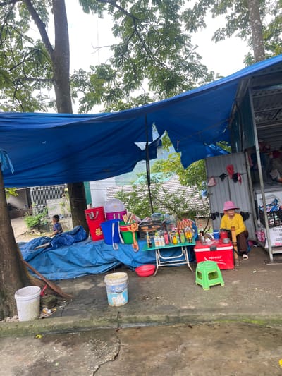 The image depicts an outdoor market stall in Lùng Tám Commune, Vietnam, situated beneath a large blue tarp supported by trees and a makeshift structure. A woman wearing a yellow shirt and a pink hat is seated on a stool, facing a table laden with various items. These include bottles of drinks, eggs in crates, and other merchandise. To the left of the stall, a young boy in a blue and checkered shirt is visible near a pile of blue tarps. Several buckets and containers, some red and blue, are arranged near the stall, along with a broom leaning against a tree. The ground is a wet, cracked concrete surface. In the background, houses with tiled roofs and lush greenery suggest a semi-rural or suburban setting. The overall scene captures a typical day at a local market.