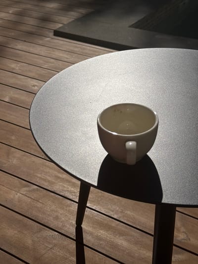 A close-up, outdoor shot focuses on a small, white ceramic teacup resting on a dark gray, circular table. The table's textured surface is illuminated by strong, angled sunlight, casting a distinct shadow of the teacup to its right. The teacup itself appears empty and shows faint brown residue on its interior. The table is positioned on a wooden deck with planks running horizontally across the frame, also patterned with shadows and highlights from the sunlight. In the background, a dark, rectangular structure, possibly a pool or a raised platform, is visible, further in shadow. The scene is devoid of any people. The image captures a moment of quiet solitude on a sunlit day, likely in a residential outdoor area.
