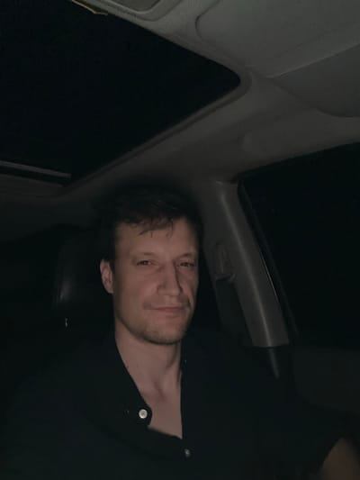 The image shows a close-up, angled shot of a man in what appears to be the interior of a car at night. The man is wearing a black collared shirt. His face is partially visible, with his eyes looking towards the camera and a slight smile. The background is dark, with some visible car interior elements like a sunroof and part of a seat. The lighting suggests it is nighttime, and the environment is indoors. There is no visible text in the image. The overall mood is casual and personal.