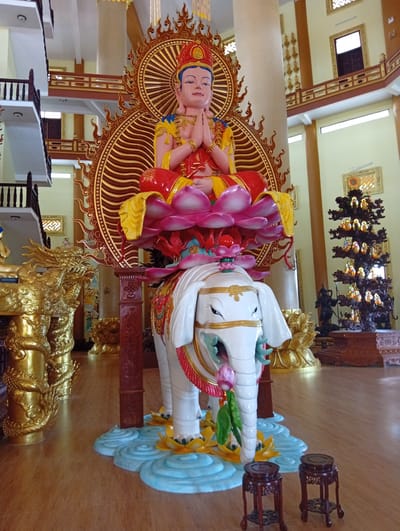 This image depicts the interior of a multi-level temple or pagoda located in Thiện Chí, Vietnam. The central subject is a large, colorful, multi-component statue prominently positioned in the center.
The primary statue features a deity, possibly a Bodhisattva, seated cross-legged atop a large pink lotus blossom. The deity wears a red and yellow robe, gold jewelry, and a red and gold crown, with hands pressed together in a prayer gesture (anjali mudra). A large, ornate golden mandorla with radiating patterns and flame-like edges frames the deity from behind. This entire upper section is supported by a large, white elephant statue adorned with gold trim, red and green accents, and a decorative saddle. The elephant holds a pink lotus flower in its trunk and stands on a light blue base designed to resemble water, featuring yellow lotus flowers.
To the left of the central statue, a large, intricately carved golden dragon statue is partially visible, coiled around a vertical structure. To the right, a dark, multi-tiered tree-like structure, possibly carved wood, displays numerous small, golden Buddha figures on its branches. A dark statue on a pedestal is visible further back on the right. In the foreground, two small, dark brown wooden stools with carved legs rest on the polished light brown wooden floor.
The background reveals a spacious interior with light cream-colored walls adorned with gold decorative motifs and framed artworks. Large white pillars support the structure, and multiple levels are visible with dark wooden railings forming balconies. Windows on the upper levels provide ambient light. Hanging decorative elements are visible from the ceiling.