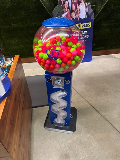 In Schaumburg, United States of America, a Topgolf branded gumball machine filled with bright pink and neon green golf balls stands on a grey concrete floor. The machine has a blue plastic base with a spiral dispenser for the golf balls and a slot for tokens, with a sign reading "TOKENS ONLY" above it. A blue and yellow sign with "FUN PASS" is visible in the background, along with artificial turf and a partial view of people. To the left, a wooden counter is visible with a blue and white stand on it. The image is taken at eye level.