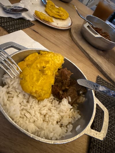 The image shows a partially eaten meal of white rice, stewed meat, and fried plantains (possibly tostones) served in a small metal pot-like container on a wooden table. A fork and knife are positioned around the pot. In the background, another similar pot is visible, along with a glass of juice. The scene likely depicts a dining setting in Punta Cana, Dominican Republic, where this type of meal is common.
