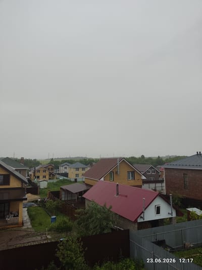 This image captures a view of a residential area in Kuyuki, Russia, under an overcast sky, suggesting a cloudy or possibly rainy day. The scene is dominated by a line of houses with varying architectural styles and roof colors, predominantly red and brown. The houses appear to be detached or semi-detached, with some featuring brick exteriors and others painted or clad in different materials. Many of the houses have visible chimneys.

The setting is a suburban or rural landscape, with greenery in the form of trees and grass surrounding the properties. Some properties have fences, and one shows a blue barrel in the yard. Power lines are visible in the distance, indicating the presence of infrastructure. The time of day is indicated by the text overlay: "23.06.2026 12:17," placing the image in the early afternoon of June 23, 2026. There are no people visible in the image. The overall mood is calm and somewhat subdued due to the overcast weather.