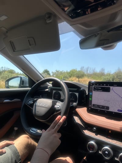 The image shows the interior of a car, a Skoda, on a sunny day, with the view looking towards the dashboard and out the windshield. A person's hands are visible in the foreground, one hand resting on the steering wheel, and the other making a peace sign. The car's digital dashboard displays navigation information for a route that includes the city of Πεντάπολη, Greece. The navigation screen shows the current time as 14:26 and indicates a distance of 11 km to the next destination, which is labeled "Περιφ. Σερρών". The car appears to be in motion, as suggested by the road and scenery visible through the windshield, and the navigation system being active. The interior of the car features brown leather trim on the dashboard and seats. The overall scene suggests a journey through a rural or suburban area, possibly during the summer months due to the bright sunlight and clear sky.