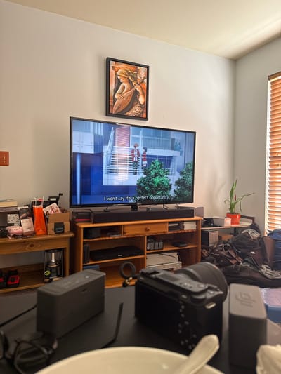 This image captures a section of an indoor room, likely a living area, bathed in natural light from a window fitted with wooden blinds at the top. The walls are a light, neutral color.
A large flat-screen television, identified by "SAMSUNG" text on its bezel, is displaying an animated program that features a multi-story building and a character on a staircase. Subtitles on the screen read: "I won't say it's a perfect opportunity." Below the television, a black soundbar is mounted. To the left of the TV, a framed piece of art, depicting an antique-style scene with a figure and a large fish or sea creature, hangs on the wall.
To the right of the television, a wooden shelving unit is filled with various objects. The top shelf holds bags, possibly luggage, and a potted plant with red flowers. Middle shelves contain electronics, including a camera with a lens, a black box labeled "DJI," and a "PHILIPS" branded DVD player. Lower shelves store boxes, one marked "prime," shopping bags, and other items, such as a "DURACELL" lantern. In the immediate foreground on the right, a white bowl or plate is partially visible, alongside additional electronic devices, one of which also bears the "DJI" logo.
The presence of natural light suggests it is daytime. The room appears lived-in, consistent with a residential setting in Glen Burnie, United States of America.