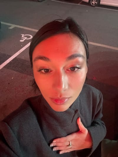 A woman in a gray coat lies on what appears to be asphalt. Her head is angled towards the camera, with a slight upward tilt. She has dark hair and fair skin, accentuated by a red light source. Her hand, adorned with rings, rests on the coat. The background shows a disabled parking symbol, hinting at the setting. The overall composition conveys a sense of casual repose and perspective.