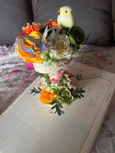 The image depicts an arrangement of artificial flowers in a decorative basket. The basket contains roses in shades of green, cream, and orange, accented with small white and pink flowers and greenery. A toy bird and decorative butterfly are also visible within the arrangement. The basket rests on a white tray with children's doodles (hearts) and writing (e.g., "Dedburg") on it. The scene is set on a patterned bedspread with floral motifs. To the left of the arrangement is a grey textured cushion. There is no indication of the city of Gorun, Bulgaria, or any people.