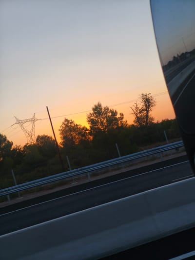 The image depicts a roadside scene at sunset, likely in Riba-roja de Túria, Spain. The sky exhibits a gradient of pale blue to warm orange hues, indicative of twilight. Silhouetted against the vibrant sky are trees and a large electricity pylon. The foreground features a road with white lane markings, separated from the embankment by a metal guardrail. The right side of the image shows a reflection, possibly from a vehicle's mirror or window, revealing a glimpse of the road and surrounding landscape. The overall impression is one of a serene, natural setting juxtaposed with human infrastructure.