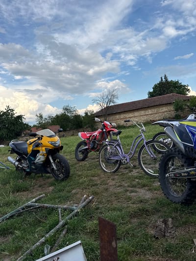 This is a full, eye-level shot of a rural yard in Bratya Daskalovi, Bulgaria, under a partly cloudy sky. In the foreground, a yellow and black motorcycle is parked on the grass, facing left. To its right, a red dirt bike is angled towards the viewer, with a purple bicycle positioned between it and another motorcycle. The third motorcycle, partially visible on the far right, is blue and white. In the background, a long, stone-built house with a red-tiled roof is visible, surrounded by trees and other vegetation. Debris, including metal poles and what appear to be old boots, are scattered across the grassy terrain.