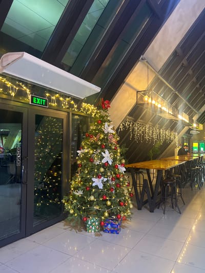 This image depicts an interior commercial space, likely a cafe or lounge, situated in Siktarva, Georgia, during the evening. A decorated artificial Christmas tree stands prominently near glass double doors on the left, adorned with warm white string lights, red and gold spherical ornaments, and white poinsettia-like decorations. Two wrapped gift boxes, one green and one blue, are positioned at the tree's base. Above the glass doors, an illuminated green "EXIT" sign is visible, topped by a white horizontal air curtain unit and draped with yellow string lights. To the right, a long, narrow wooden table with dark metal "W"-shaped legs extends along the wall, accompanied by multiple dark metal bar stools. Overhead, linear black metal light fixtures with exposed bulbs hang from an angled, dark-paneled ceiling, below which more yellow string lights are draped in a cascaded pattern. One male individual, wearing a dark top and possessing dark hair, is seated at the far end of the long table, facing away from the camera, appearing to interact with an electronic device. The floor consists of light-colored tiles, reflecting the ambient lighting.