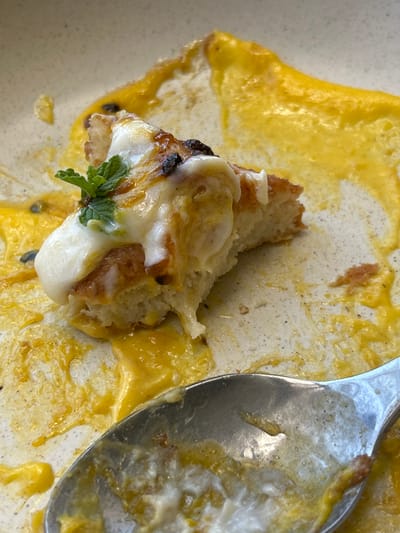 This close-up image depicts a partially consumed dessert or breakfast item on a light-colored, speckled ceramic plate. A substantial piece of a light-brown, bready item, possibly a type of French toast or pastry, is positioned centrally. It is topped with a melted white cream or cheese and a small cluster of fresh green mint leaves. Darker, caramelized portions are visible on the bready item's surface. The plate is extensively smeared with a viscous, bright yellow-orange sauce or fruit puree, containing small dark specks consistent with passion fruit seeds. A silver-colored spoon, appearing used and stained with remnants of the white cream and yellow sauce, is visible in the bottom right corner of the frame. The scene suggests a dining environment, possibly a restaurant or cafe, in the city of El Tezal, Mexico. No individuals are present in the image.