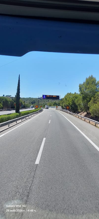 The image shows a road in Calafell, Spain, with a sign indicating "ZONA DE OBRAS" (Construction Zone). The road is paved with asphalt and has a white line marking the lanes. There is a metal guardrail along the side of the road. The sky is blue, and there are trees and vegetation alongside the road. A tall, slender tree is visible. The image was taken from inside a vehicle, as evidenced by the car's interior frame in the left side of the photo. The timestamp indicates the photo was taken on May 26, 2026, at 14:48. A speed limit sign with "50" is visible.