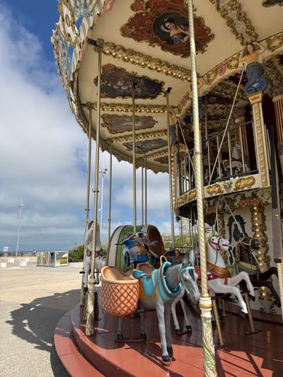 An outdoor carousel in Berck, France, is depicted with a partial view of its ornate canopy, adorned with paintings and decorative lights. The carousel features several horses and one grey donkey, each with detailed saddles and bridles. The foreground shows the wooden platform of the carousel, with shadows cast on the surrounding paved area. In the background, a street with bicycles and a few people is visible, along with a cloudy sky. The carousel appears to be situated near a beach or promenade, indicated by the expanse of paving stones and a glimpse of what might be the sea or a body of water in the distance.