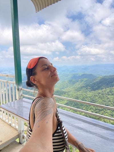 A woman with an orange eye mask on her forehead is standing on a balcony overlooking a lush green mountain range. The sky is blue with scattered clouds. She is wearing a black and white striped top. There are tattoos on her arm and shoulder. A table with a marble top is visible behind her, along with white railings. The location is identified as Phan Son, Vietnam.