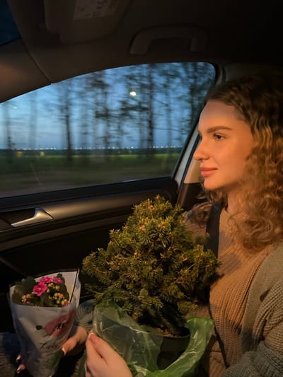 A woman with curly blonde hair is inside a car, looking down at a small coniferous tree in a plastic bag and a bouquet of pink flowers in a paper wrapper. She wears a tan sweater and a dark seatbelt. The car window shows a blurred view of trees and a twilight sky. The scene takes place at night or dusk in Дубровка, Russia. The interior of the car and the objects held by the woman are visible.
