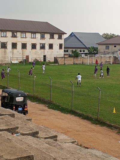 The image depicts an outdoor football match in Ejisu, Ghana, observed from elevated concrete spectator stands. In the immediate foreground, weathered grey concrete steps of the bleachers are visible, with some refuse present on their surfaces. Below the stands, a reddish-brown dirt path runs horizontally across the frame. A black three-wheeled vehicle, resembling an auto-rickshaw, is parked on this path, displaying the license plate "AW 85-44 ASH" and the text "Amanfuo Hyia" on its rear window. A chain-link fence, supported by vertical metal posts, separates the dirt path from a green grass football pitch.
On the football pitch, approximately 14-15 male individuals are actively engaged in play. They wear two distinct uniform colors: white and purple/lavender. One player in a white jersey with the number "13" on his back is positioned near the center of the field. A player in a red jersey, presumed to be a goalkeeper, is visible near the goal frame on the right side of the field, which has a white net. Another player in a purple jersey is captured in motion, appearing to dribble or kick a white football near the left mid-field. A second metal goal frame is partially visible on the left side of the field.
In the background, behind the football pitch, stands a large, multi-story building with a brown tiled roof, light-colored, weathered walls, and multiple windows fitted with security bars. To the right of this structure, a building with a dark grey, steeply pitched roof is visible, along with parts of another light-colored building further to the right. A low concrete block wall separates the playing field from these background structures. The sky above is a uniform, light grey, indicating overcast conditions or a hazy atmosphere. A single yellow traffic cone is positioned on the dirt path near the right edge of the frame.
