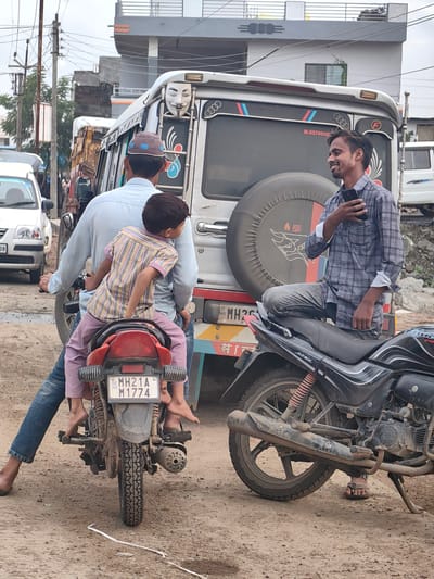 In Padli, India, a man wearing a checkered shirt and jeans stands next to a parked motorcycle, holding a smartphone. He is looking to his right and smiling. Behind him, a man in a light blue shirt and a child in a striped shirt are seated on another motorcycle, facing away from the camera. The child is looking to the right, and the man appears to be looking forward. A large vehicle, possibly a bus or van, is parked behind them, decorated with a Guy Fawkes mask graphic on its side. The scene takes place on a dirt road with other vehicles and buildings visible in the background. The sky is overcast.