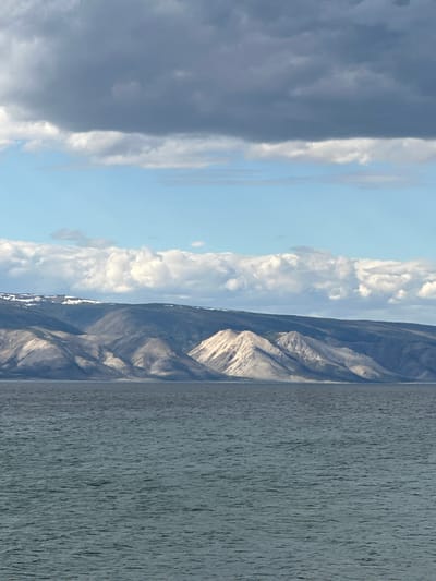 The image depicts a landscape with a body of water, mountains, and sky. A body of water dominates the right side, its surface appearing textured and slightly disturbed, possibly by wind. Adjacent to the water, a mountain range rises, its slopes and peaks marked by varied shades of grey and hints of white, possibly indicating snow. The sky occupies the left portion of the image, featuring a mix of dark, heavy clouds on the left and lighter, more scattered clouds towards the center. Between these, there's a patch of blue sky. The photograph shows an image of Lake Baikal in Khuzhir, Russia.