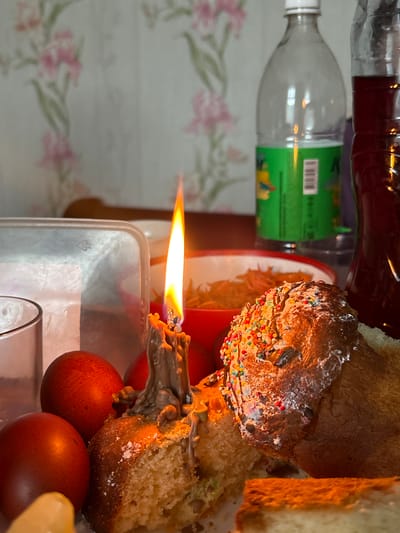 The image depicts a celebratory scene, possibly Easter. A partially sliced and decorated sweet bread (Kulich) sits atop a plate, alongside dyed red eggs. A lit candle is placed among the Kulich and eggs. Glass bottles are present in the background, along with a floral patterned wall. The location, Пожарское сельское поселение, Ukraine, is provided.