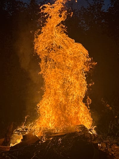 A large bonfire burns intensely in the darkness, with flames reaching high into the night sky. The fire is primarily orange and yellow, with wisps of smoke curling upwards. The base of the fire appears to be built upon wood and other combustible materials, including what looks like branches and debris. In the background, the dark silhouettes of trees are visible against a dark sky. The scene is illuminated by the bright glow of the fire. There is no indication of the location within the city of Rasskazovo, Russia, nor are there any people or specific actions depicted.