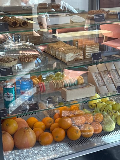 The image shows a display case filled with various food items, including pastries, sandwiches, macarons, fruits, and bottled drinks, likely in a bakery or cafe. A glass partition separates the viewer from the merchandise. There are price tags visible in front of some items. The lighting is artificial, creating reflections on the glass. In the background, blurred figures and furniture suggest an indoor commercial setting. The specific location in Patrusheva, Russia, is not discernible from the image itself, but the presence of Russian language on some labels confirms a Russian-speaking context.