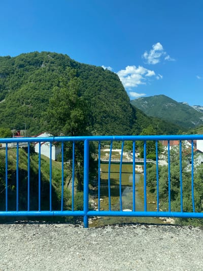 The image shows a view from a bridge railing in Šavnik, Montenegro. The railing is blue and made of metal bars. In the foreground, the bridge's concrete surface is visible, covered in small pebbles. Beyond the railing, a river flows through a valley with steep, tree-covered mountains in the background. Several buildings, including a white one with a red roof, are visible along the riverbank. A smaller bridge crosses the river in the distance. The sky is clear blue with a few white clouds.