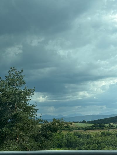 The image depicts a landscape with a dramatic sky filled with dark, overcast clouds. In the foreground, a large evergreen tree with dense foliage dominates the left side of the frame. Beyond the tree, rolling hills covered in green vegetation stretch into the distance. A distant range of mountains can be seen on the horizon, appearing hazy and muted by the atmospheric conditions. The overall impression is one of a natural, rural setting under a brooding sky, potentially before or during a rain shower. There are no people or man-made structures clearly visible in the scene, apart from what appears to be a railing in the very bottom of the frame. The location is identified as Rajana, Bulgaria.