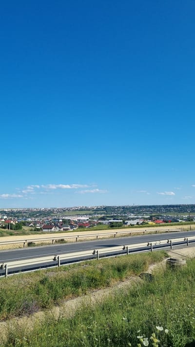This is a photograph taken from an elevated position, looking towards a town under a clear blue sky with a few scattered white clouds. In the foreground, a section of a highway is visible with a beige guardrail. The highway is empty. Beyond the highway, the landscape slopes gently upward, revealing a residential area with many houses with red-tiled roofs. Further in the distance, the town extends, with commercial buildings and more houses scattered across the hills. The overall scene suggests a bright, sunny day in a suburban or semi-rural area. The location is the outskirts of Șcheia, Romania, with the highway likely being the DN2, a major road connecting Bucharest to the northern part of the country, and the town of Șcheia being a commune in Suceava County.