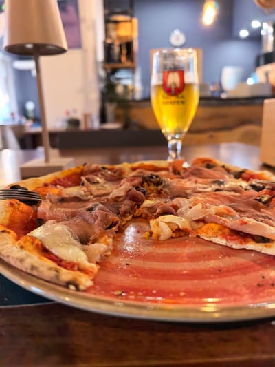 A close-up shot captures a half-eaten pizza on a red and white striped plate, resting on a wooden table. The pizza features a crust with char marks, topped with tomato sauce, melted mozzarella cheese, thinly sliced prosciutto, and black olives. A fork is partially visible on the left side of the plate, indicating the pizza is being eaten.
In the background, a glass of light golden beer with a frothy head is positioned behind the pizza. The beer glass has a crest with a red shield and a white emblem, along with some text beneath it that is partially obscured. The setting appears to be a restaurant or bar, with warm lighting emanating from a lamp on the left and overhead lights in the distance. Out-of-focus shelves with bottles and other items are visible in the background, suggesting a casual dining environment. The overall mood is relaxed and inviting, capturing a moment of enjoying a meal.
The time of day is likely evening or late afternoon due to the warm artificial lighting. There are no obvious weather cues. The location context of San Giovanni in Persiceto, Italy, suggests a European dining experience. There is no visible text other than that on the beer glass, which is largely illegible.