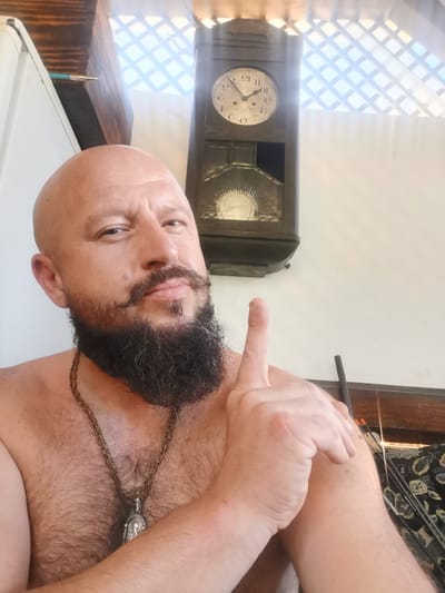 A bald, bearded man with a mustache is shown from the chest up, looking towards the camera. He is pointing his right index finger upwards, with his arms crossed in front of him. He is wearing a chain necklace with a pendant. In the background, a vintage clock is visible on the wall. The image appears to have been taken indoors. There is no indication of the location in the city of Semypolky, Ukraine.