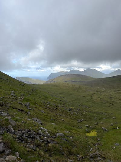 An outdoor, vertical, wide-angle shot depicts a lush, green, mountainous landscape under a heavily overcast sky. The foreground is dominated by a steep, grassy slope littered with rocks and patches of moss. A faint, rocky path is visible winding its way down the slope from the left.
In the midground, rolling hills covered in vibrant green grass stretch towards the horizon. These hills are textured with subtle undulations and the occasional small watercourse. A distinctive, flat-topped cliff formation juts out into the sea on the left side of the image, with smaller cliffs visible in the distance on the right.
The background consists of several layered mountain ranges, their peaks softened by the atmospheric haze and the thick cloud cover. The sea is visible between the prominent cliff formations, appearing calm under the diffused light. The sky is filled with heavy, grey clouds, with hints of lighter patches suggesting the presence of the sun behind them.
The overall impression is one of rugged natural beauty, characterized by vast expanses of green, dramatic coastal features, and a subdued, moody atmosphere typical of a cloudy day in a northern climate. The scene is devoid of any human presence or man-made structures, emphasizing the wildness of the environment. The location is the Faroe Islands, specifically Tjørnuvík, a village known for its dramatic natural scenery.