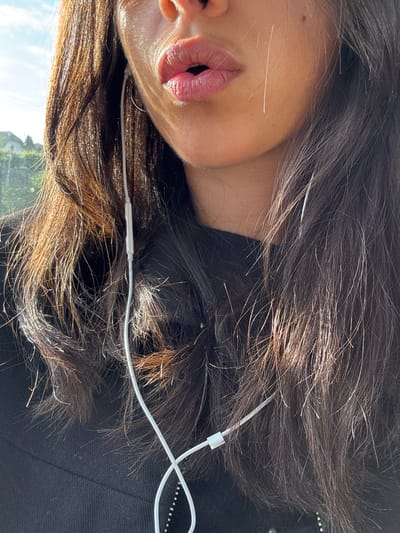 The image presents a tight, vertical close-up of a person's face from approximately the nose down, encompassing the mouth, chin, jawline, and a significant portion of dark hair. The individual has medium-toned skin, illuminated by strong, warm sunlight that creates distinct highlights on the skin, lips, and hair. Their lips are slightly parted in an 'O' shape, suggesting they might be speaking or singing, and appear to have a natural pink or subtle lip tint.
Long, dark brown hair cascades around the face and shoulders, with numerous strands illuminated by the sun, showcasing lighter brown or reddish highlights and individual hairs. A white wired earbud cord is prominent, draped over the left ear (from the viewer's perspective), extending downwards across the chest. A rectangular inline remote/microphone unit is visible on the cord near the left ear. A thin, metallic hoop earring is visible in the left ear. A small, dark mole or freckle is present on the right cheek, slightly below the nose. The individual wears a black garment, likely a shirt or sweater, visible on the lower left side of the frame.
The background is out of focus, revealing a bright outdoor environment. In the upper left, a segment of blue sky with faint white clouds is visible. Below the sky, blurred green foliage and a distant, indistinct light-colored building with a dark roofline are discernable. The image does not contain any specific architectural features, landmarks, or textual information that would allow for the identification of the location as Baccarat, France, or any other specific city.