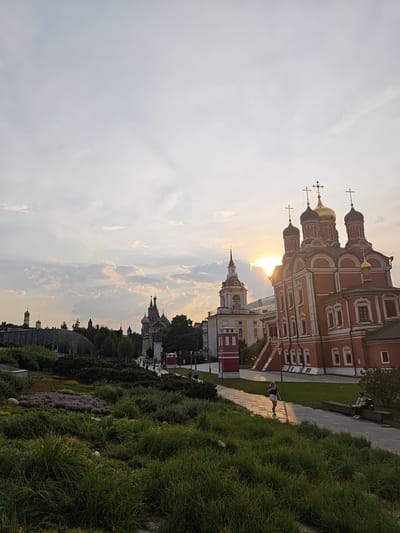 The image captures a sunset scene in a city, likely Moscow, given the architectural style of the churches and the presence of many domes, which are characteristic of Russian Orthodox churches. In the foreground, a lush green landscape with manicured bushes and flowers leads to a pathway. Several people are visible on the pathway and sitting on a bench. To the right, a vibrant red church with golden domes stands prominently. The sun is setting behind the church, casting a warm glow and dramatic light. In the background, more churches and buildings with similar architectural features are visible against a cloudy sky. The overall impression is one of serene beauty and architectural grandeur, typical of a historic city center. The location is not Путрышкаўскі сельскі Савет, Belarus.