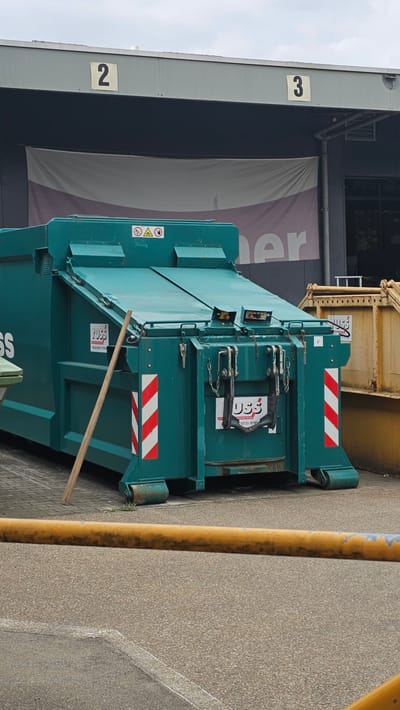 An outdoor, medium-shot, eye-level photograph captures a large, dark teal industrial waste bin with a hinged lid and an attached compactor, positioned on a paved surface. The bin, manufactured by "FUSS," is marked with red and white reflective stripes on its rear corners, and bears a "FUSS" logo with a phone number. A wooden pole leans against the side of the bin. In the background, a grey industrial building features prominent numbered signage: "2" on the left and "3" on the right. A large banner, partially visible, hangs across the building's facade. To the right of the teal bin, a portion of a yellow industrial container is visible. A yellow horizontal barrier runs across the lower foreground. The scene appears to be located within an industrial area of Neu-Ulm, Germany, indicated by the building's architecture and the presence of industrial equipment.