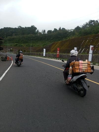 This image depicts an overcast daytime scene on a paved road in Saing Ta Mawt Lay, Myanmar, showing two motorcycles traveling away from the viewer.
In the foreground, a person wearing a white full-face helmet and dark clothing operates a grey scooter heavily laden with cargo. A large, light-brown wooden crate secured to the rear rack contains numerous individually packaged items, predominantly in pink and white wrappers, suggesting confectionery or snacks. Additional packages, including a bulky brown bag, are strapped to the left side of the scooter. The operator's bare feet are visible, indicating they are wearing sandals or no footwear.
Approximately 15-20 meters ahead, a second person on a darker colored scooter also travels away from the viewer. This rider wears a helmet and carries a brown backpack. A barefoot passenger, dressed in dark pants, is seated behind the rider.
The road surface is dark asphalt, delineated by a solid white line on the left shoulder and a double solid yellow line in the center.
To the right of the road, a dark grey stone retaining wall, partially covered in green moss, supports a steep, vegetated hillside. Several vertical banners are affixed to the wall: two plain white, one red with partially visible text (e.g., "SLOP MANG"), and a white rectangular board displaying a red circular "No Parking" symbol. Power lines are visible in the upper background above the hillside.
To the left of the road, a dark architectural structure is visible near the shoulder, accompanied by two orange traffic cones. Beyond this, a dense forest of green trees and undergrowth extends upwards. The sky is uniformly light grey and overcast.