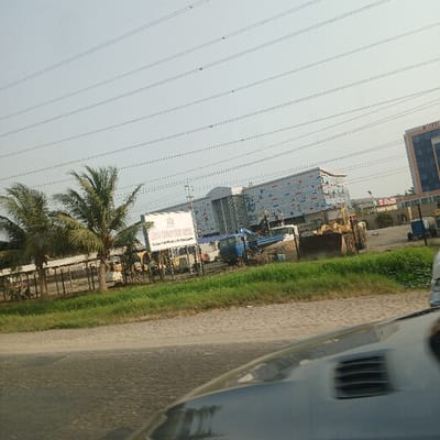 The image depicts a bright, sunlit outdoor scene, captured from a moving vehicle looking towards the side of a road. In the immediate foreground on the right, a portion of a dark-colored vehicle, likely a car, is visible, including a vent or grille.
A paved road stretches from the foreground into the distance, bordered by a green grassy median strip. Several lush palm trees are scattered along this median and the adjacent areas, suggesting a warm climate.
The midground and background showcase an active urban or semi-urban development. A large construction site is evident, populated with numerous yellow construction vehicles, including what appear to be bulldozers and excavators, and a blue crane-like machine. Behind this site, several buildings are under construction or recently completed. One prominent building features a modern facade with a grid pattern of blue and light-colored squares. Further in the background, a taller, darker building, possibly a high-rise, can be seen.
A large white billboard is visible near the construction area. Partially legible text on it reads "CHINA CONSTR...", suggesting a Chinese construction company or project. Overhead, numerous power lines crisscross the clear, pale blue sky.
The time of day appears to be midday due to the bright, clear conditions. The scene conveys a sense of ongoing development and construction activity in a location consistent with a road in Nigeria.
