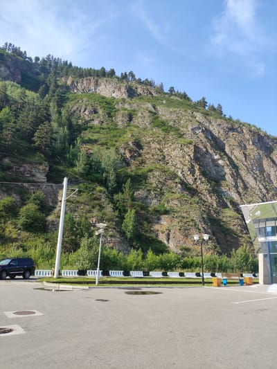 This image captures a scenic view of a rocky mountain slope covered in lush green vegetation and scattered trees. The sky above is a clear, bright blue with a few wisps of white clouds. In the foreground, there's a paved area, likely a parking lot, with puddles of water indicating recent rain. A black SUV is parked on the left side of the frame. A white utility pole with wires stretches up the slope, and a lamppost stands near a bench with blue trash receptacles. To the right, a modern building with a reflective glass facade is partially visible. The overall scene suggests a natural landscape combined with man-made infrastructure, possibly a rest stop or a viewpoint. The lighting indicates it is daytime, likely late morning or early afternoon, given the bright sunlight and clear sky. The environment appears calm and serene. There is no visible text in the image.