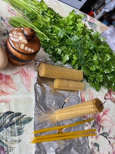 An overhead, close-up shot shows various items arranged on a tablecloth with a floral pattern. In the upper left, a bundle of green herbs, possibly dill and parsley, rests next to a wooden jar with a decorative lid. To the left of the herbs, an egg is partially visible. In the center of the image, two honeycomb beeswax candles are positioned on aluminum foil. A third, larger honeycomb candle lies on the foil, with a lit wick and some yellow wax drippings. Three thin, yellow taper candles are placed below the larger candle. The background shows a blurry glimpse of a kitchen area. The scene is likely indoors, possibly in a residential setting.