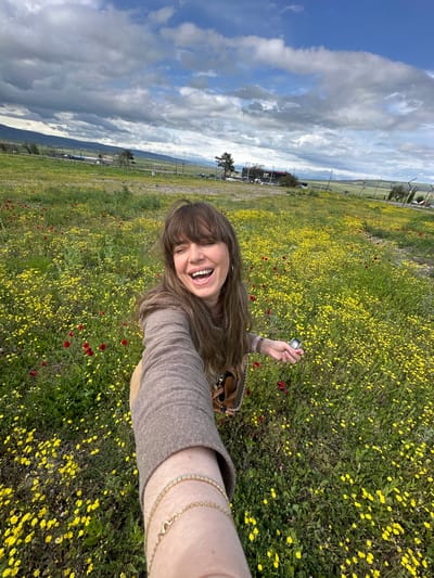 A woman is taking a selfie in a field of yellow and red flowers, under a partly cloudy sky. The location appears to be in Marneuli, Georgia, evidenced by the landscape and fence line in the background. The woman has brown hair and is wearing a tan sweater. She is smiling broadly. In her hand, she is holding a metallic object, possibly a camera. She is also wearing a gold bracelet on her wrist. The field stretches towards the horizon, and beyond the fence line, there are mountains in the distance.