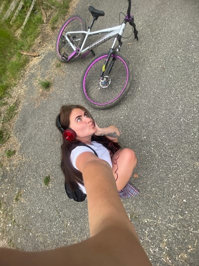In Frontoviki, Russia, a woman with long brown hair, wearing red headphones and a white t-shirt, sits on an asphalt path next to a bicycle. The bicycle is silver with purple accents on its wheels and handlebars. The woman is looking upwards, with her lips pursed, and appears to be taking a selfie. The path is bordered by grass and some dried vegetation.