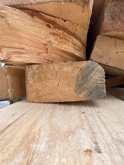 The image shows a close-up view of stacked lumber, likely at a construction site or lumberyard in Wismar, Germany. The wood is light brown and appears rough-sawn, with visible grain patterns and some darker staining on one end of a piece. The lumber is stacked horizontally, with several pieces angled in different directions. The foreground is dominated by a wide, flat piece of lumber with a textured surface. The background is out of focus but suggests more stacked wood. The overall scene is dimly lit, creating shadows and highlighting the textures of the wood. There are no people or animals visible in the image, and no activity is taking place. The environment appears to be outdoors, given the natural light and the presence of what might be a paved or gravel surface in the very bottom left corner.