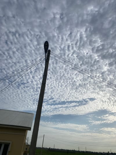 In Dumnga, Nigeria, a utility pole with a streetlamp and several wires stands against a sky filled with altocumulus clouds. The clouds are densely packed, creating a textured, rippled appearance. In the lower left of the image, a section of a yellow building with a white metal roof is visible. Power lines extend from the utility pole across the frame. In the distance, power lines and pylons are visible across a grassy plain, suggesting a rural or semi-urban setting. No people are clearly depicted in the scene. The overall impression is one of a clear day with interesting cloud formations.