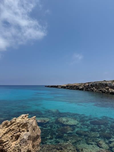 A wide-angle shot captures the stunningly clear turquoise waters of the Mediterranean Sea meeting a rocky coastline under a bright blue sky with scattered white clouds. In the foreground, a rough, sandy-colored rock formation juts out towards the water. The water itself is incredibly transparent, revealing a rocky seabed with variations in depth and color, transitioning from a lighter turquoise near the shore to a deeper blue further out. Along the right side of the frame, a rugged, light-brown and grey rocky cliff face stretches into the distance, dotted with sparse vegetation. The horizon is clearly defined, separating the calm sea from the expansive sky. This scene is characteristic of the beautiful coastal areas found in Ayia Napa, Cyprus, known for its pristine beaches and crystal-clear waters. There are no people or any discernible objects or actions in the frame.