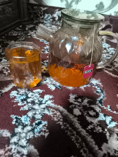 In an indoor setting in Karmana, Uzbekistan, two clear glass vessels containing amber-colored liquid are displayed on a patterned textile surface. A faceted glass teapot, approximately one-third full, holds a submerged tea bag with a pink label marked "BAYCE." Condensation is visible on the interior surfaces of the teapot and its clear lid, indicating hot contents. To the left of the teapot, a matching faceted glass cup is nearly full with the same amber liquid. Both vessels rest on a dark maroon or brown rug featuring abstract patterns in light blue, white, and beige. The background is out of focus, showing darker tones with faint lighter designs. No individuals are present.