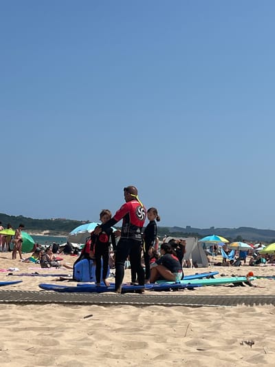 The image depicts a daytime beach scene in Ribamontán al Mar, Spain, under a clear blue sky. In the foreground, a light-colored sandy beach occupies the lower third of the frame, with a horizontal section of a patterned, flat walkway or matting visible near the bottom edge.
The midground features a group of individuals centered around what appears to be a surf or bodyboard lesson. An adult male, likely an instructor, stands prominently in a black and red full-body wetsuit, displaying a white circular, stylized spiral logo on his back and a yellow lanyard around his neck. He is addressing at least three children wearing black full-body wetsuits, who are positioned around him, one to his right and two to his left. Several blue and one teal surfboard are laid flat on the sand in front of and around this group. Multiple blue bodyboards are also present on the sand near the instructor. To the right of this central group, a young female is seated on the sand, wearing a dark top and light bottoms, observing the activity.
The background shows numerous other beachgoers scattered across the sand, engaged in recreational activities. Many are seated or lying under various beach umbrellas in colors such as green, blue, yellow, and multi-striped, or under grey pop-up shelters. Beyond the beach, a calm body of water is visible, bordered by a distant, green, tree-covered landmass or hills that extend across the horizon. A faint, possibly structural, detail can be discerned on the distant hills towards the right side of the frame.