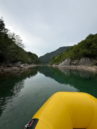 The media file depicts a scenic view from a yellow raft on a calm, clear river. The raft's yellow inflatable tubes are prominent in the foreground, with some water droplets visible on the surface. The river flows through a valley flanked by steep, tree-covered mountains. The water is a translucent green, revealing the rocky riverbed below in some areas. The reflection of the mountains and trees on the water's surface creates a mirror-like effect, adding to the serene atmosphere.

The scene is likely set in a natural, mountainous region, consistent with the provided location context of Džajići, Bosnia and Herzegovina. The time of day appears to be overcast or early morning/late evening, given the soft, diffused light and the pale sky. There are no people clearly visible in the frame, suggesting a solitary or quiet experience on the water. The overall mood conveyed is one of peace and tranquility, emphasizing the beauty of the natural landscape. The image is oriented sideways, requiring rotation for a standard view. No visible text is present in the media file.